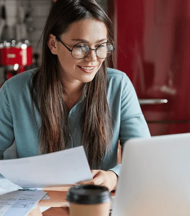 Smiling woman with glasses reviewing papers at a desk with a laptop and coffee cup