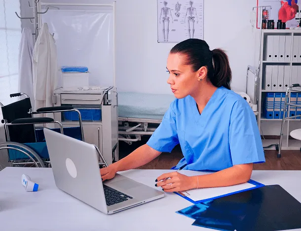 Healthcare worker in blue scrubs typing on a laptop at a desk in a clinic room