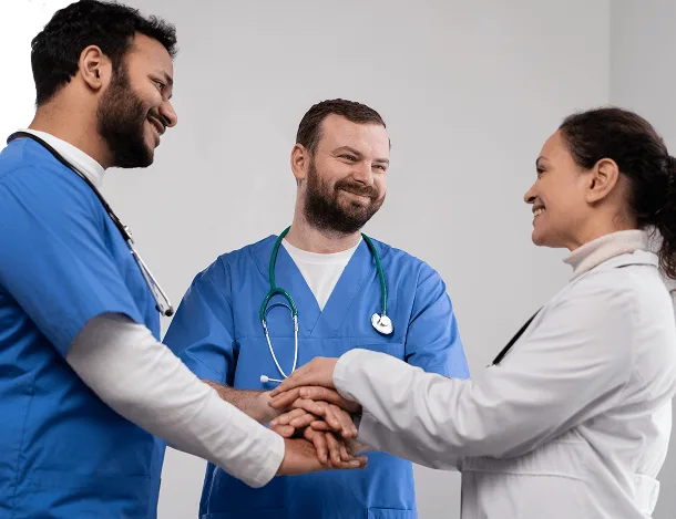 Two healthcare workers in blue scrubs and a doctor in a white coat stacking hands together
