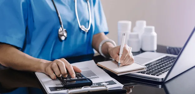Healthcare worker in blue scrubs using a calculator and writing notes beside a laptop and pill bottles