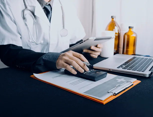 Doctor with a stethoscope using a calculator over paperwork on a clipboard, with laptop and tablet nearby