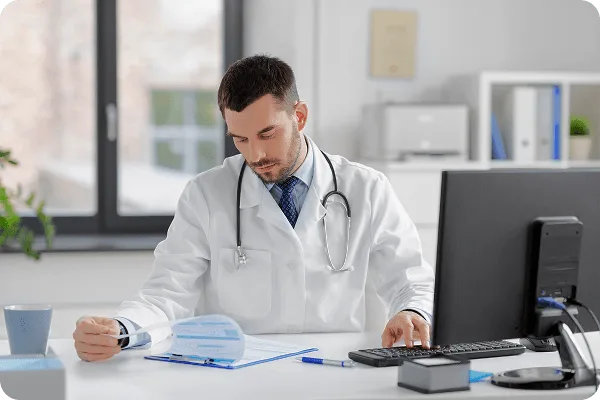 Doctor in white coat with stethoscope reviewing papers on a clipboard at a desk with a computer