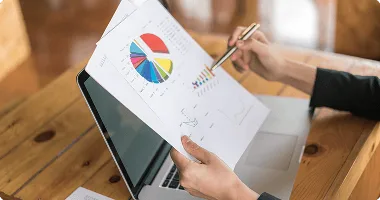 Hands holding printed pie and bar charts above an open laptop on a wooden table