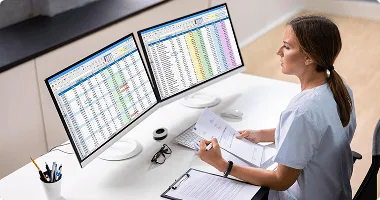 Woman in scrubs seated at a desk with two monitors showing spreadsheets, holding papers beside a clipboard