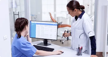 Two healthcare workers in scrubs and a lab coat looking at a computer screen in a clinic