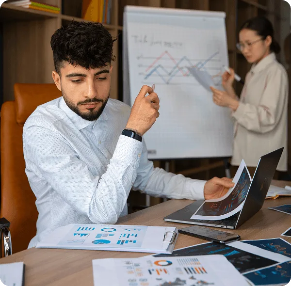 Man seated at a table with printed charts and a laptop while a woman presents a flipchart behind him