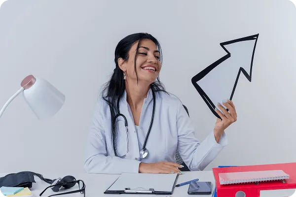 Smiling woman in white coat with stethoscope at a desk holding a large paper arrow