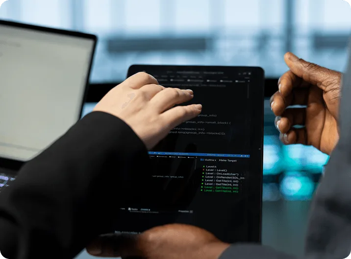 Two people pointing at code displayed on a tablet beside a laptop in a dim office