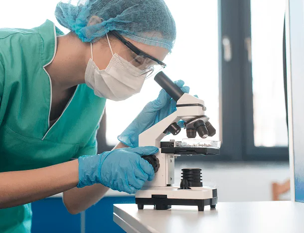 Laboratory technician in scrubs, mask and gloves looking into a microscope