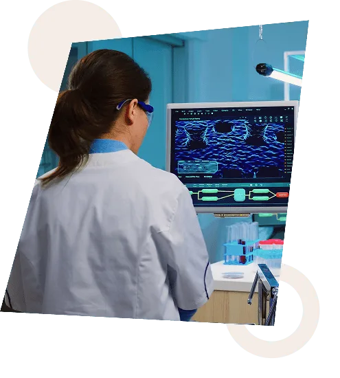 Researcher in a lab coat and safety glasses viewing data on a computer monitor