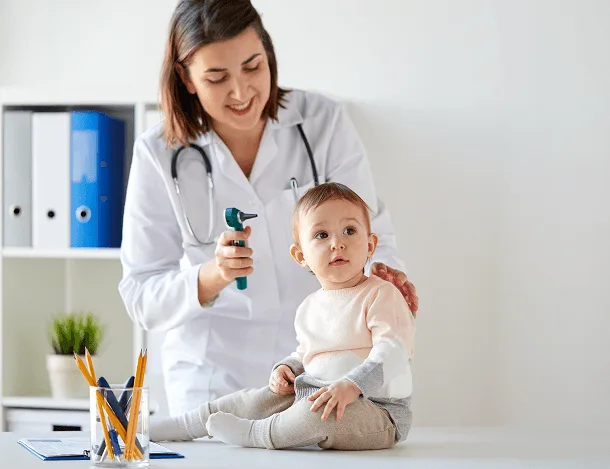 Smiling doctor holding an otoscope beside a seated infant in a bright exam room