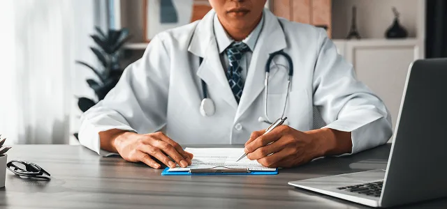 Doctor in a white coat and stethoscope writing on a clipboard at a desk beside a laptop