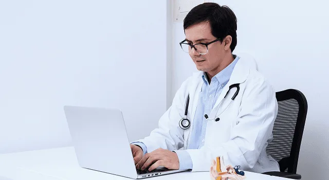 Doctor with glasses and stethoscope typing on a laptop at a white desk