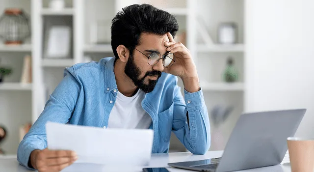 Man with glasses resting his head on his hand while reading a paper document beside a laptop