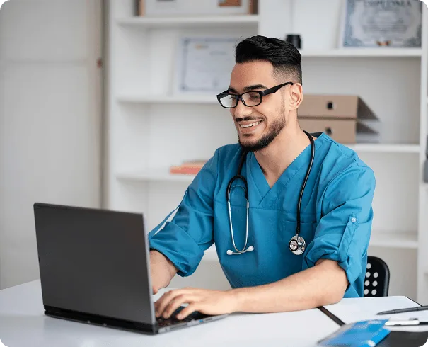 Smiling healthcare worker in blue scrubs with a stethoscope typing on a laptop at a desk