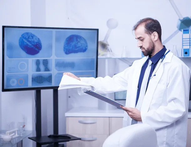 nephrology image 59705 webp Doctor in white coat reading a clipboard beside a screen showing brain scans
