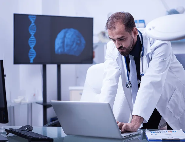 Doctor in a white coat typing on a laptop beside a screen showing a DNA strand and brain