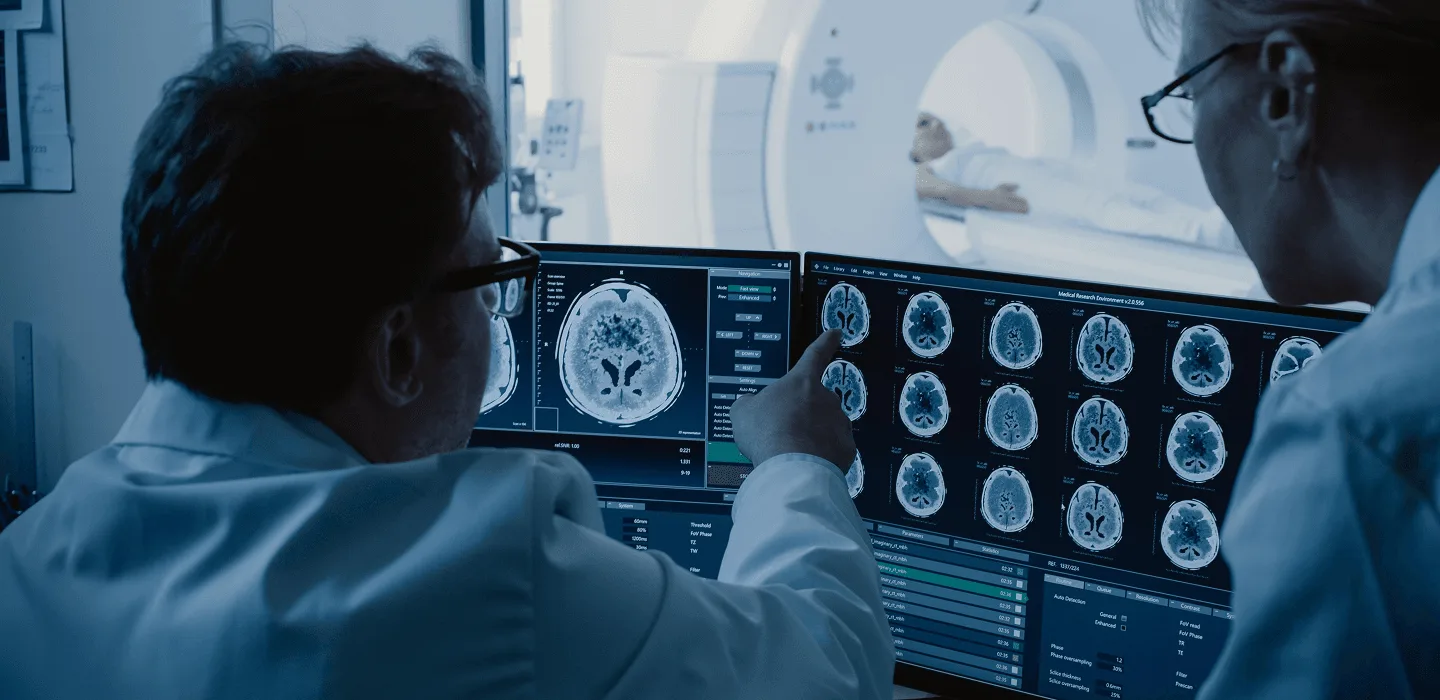 Two doctors review brain scan images on monitors while a patient lies in an MRI scanner behind them