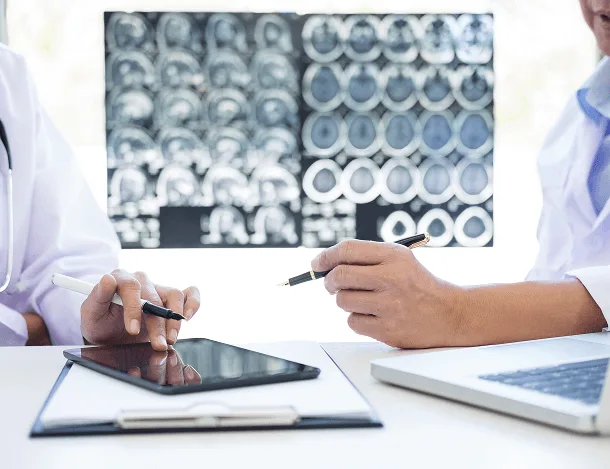 Two doctors with pens discussing printed brain scans, with a tablet and laptop on the desk
