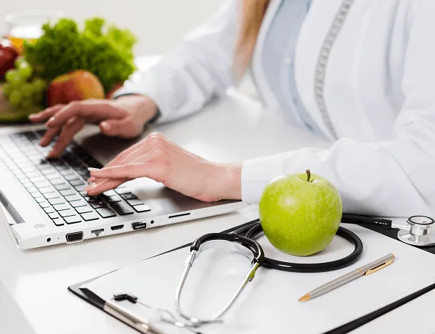 Person in white coat typing on a laptop beside an apple and stethoscope