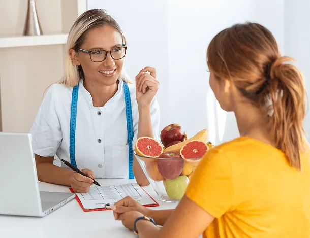 Smiling dietitian with measuring tape talks with a patient across a table with a fruit bowl