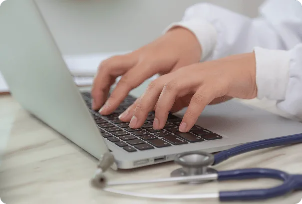 Hands typing on a laptop keyboard with a blue stethoscope resting on the desk
