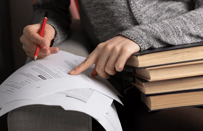 Person pointing at printed documents while holding a red pencil, with a stack of books beside them