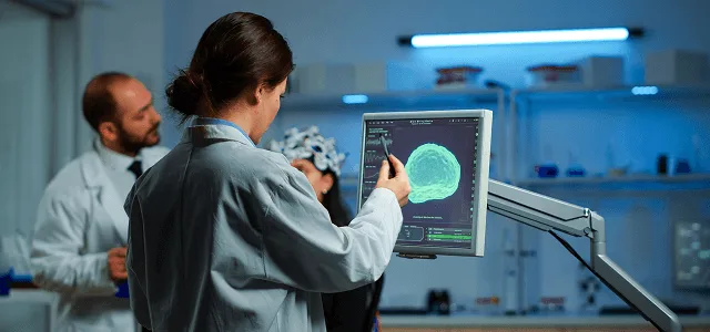 Researcher in a lab coat touches a monitor displaying a green brain scan while colleagues stand behind
