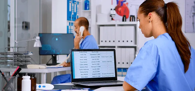 Nurse in blue scrubs reviews a patient list on a laptop while a colleague talks on the phone