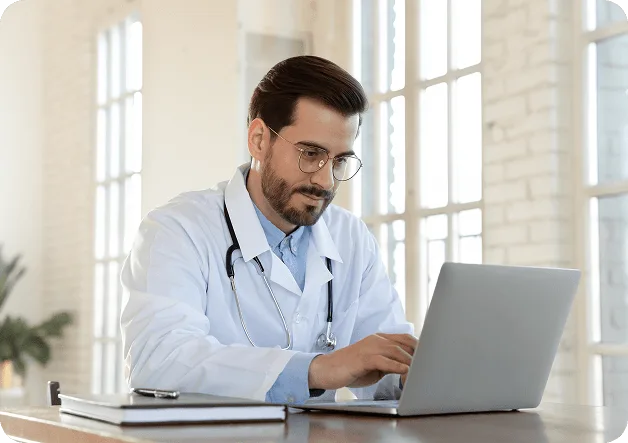 Doctor with glasses and stethoscope typing on a laptop at a desk near bright windows