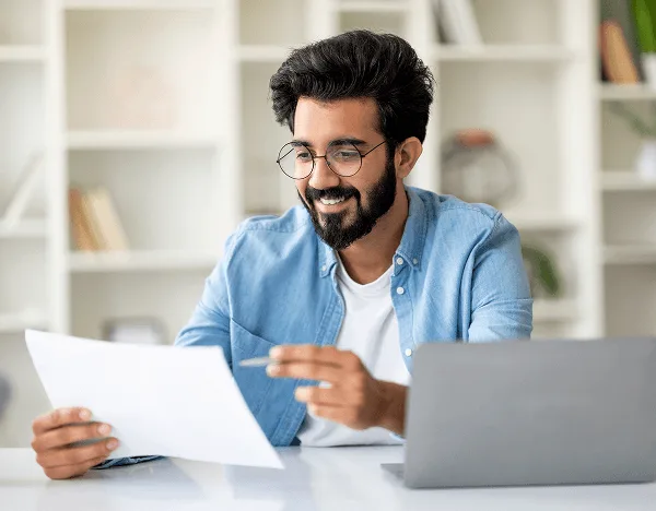 Smiling bearded man with glasses reviewing a paper document beside an open laptop