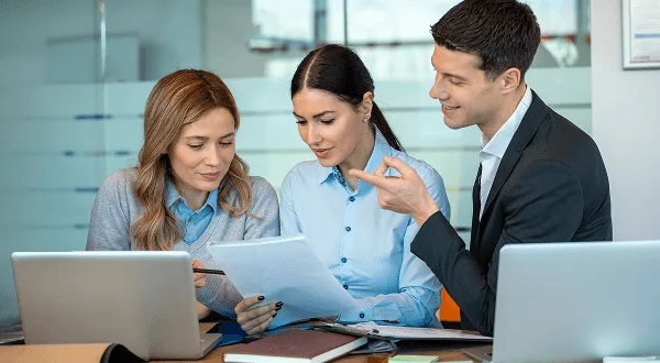 Three colleagues at a desk with laptops looking over a paper document together