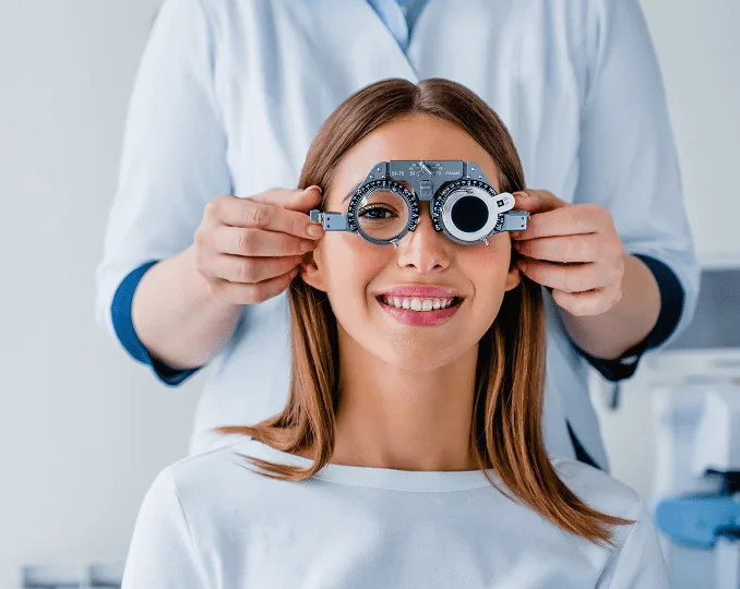 optometry image 59578 webp Smiling woman wearing a phoropter while an optometrist stands behind her