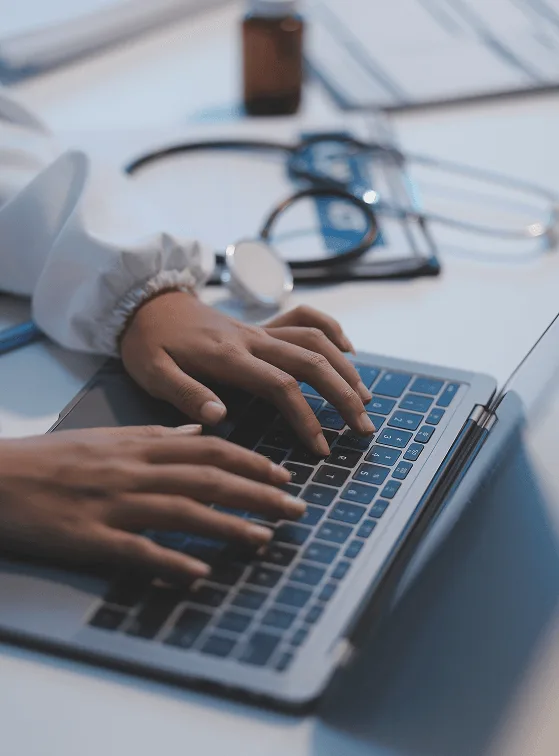 Hands typing on a laptop beside a stethoscope and medicine bottle on a desk