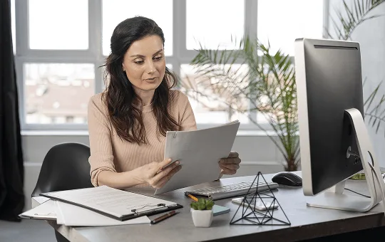 Woman at a desk reading papers beside a computer monitor and clipboard