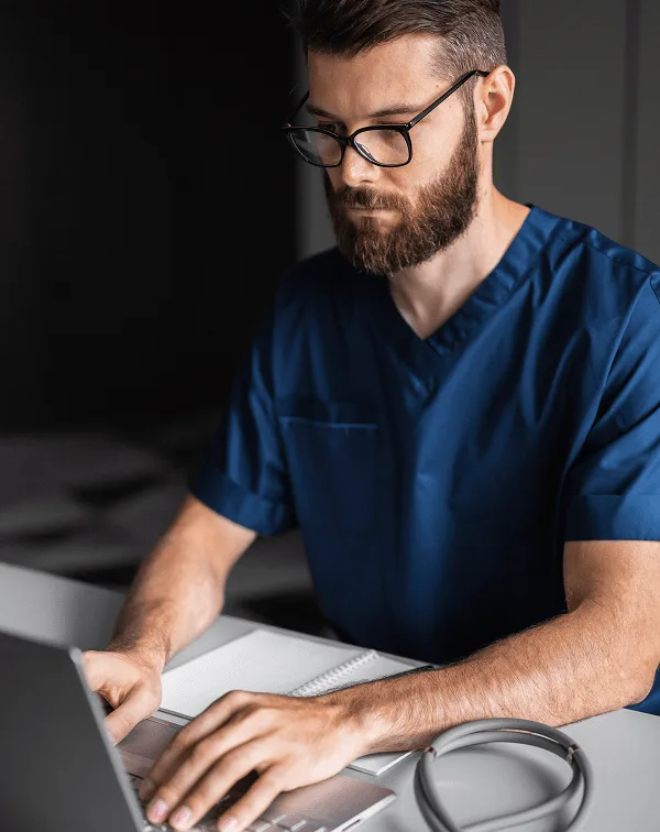 Bearded man with glasses in dark blue scrubs typing on a laptop with a stethoscope nearby