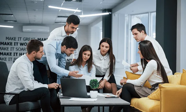 Group of office colleagues gathered around a laptop and papers on a table, discussing work