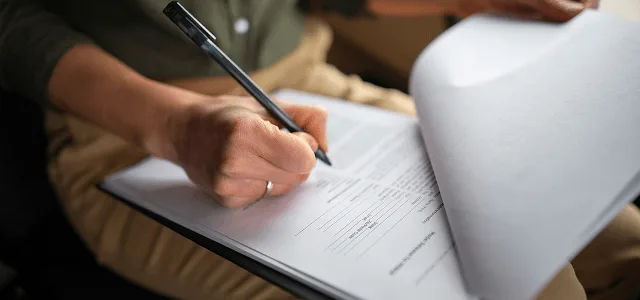 Person filling out a form on a clipboard with a pen while another page is held up