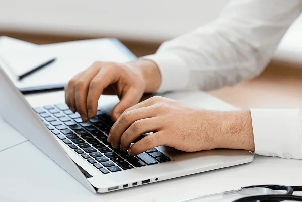 Close view of hands typing on a laptop keyboard with a pen and notebook nearby
