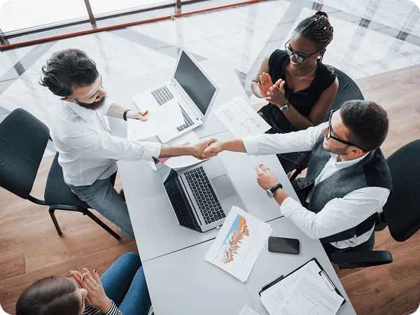 Group of colleagues from above shaking hands across a desk with laptops and printed charts