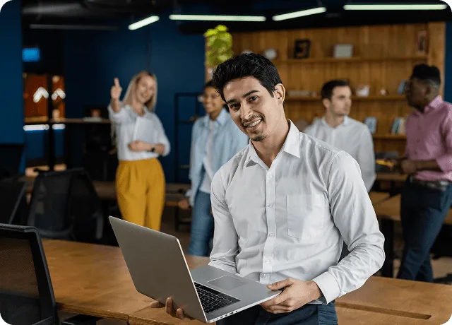 Smiling man with a laptop in a modern office while colleagues talk in the background