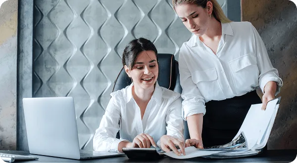 Two women in white blouses reviewing documents at a desk with a laptop, one smiling