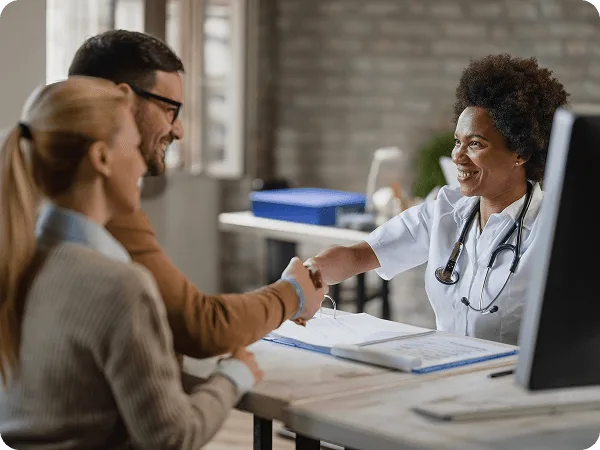 patient-statements image 60592 webp Smiling doctor shaking hands with a man at a desk while a woman sits beside him