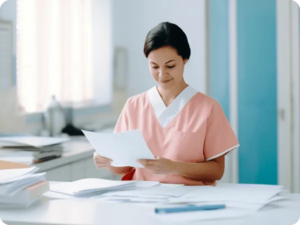 patient-statements image 60602 webp Woman in pink scrubs reading a sheet of paper at a desk covered with stacks of documents