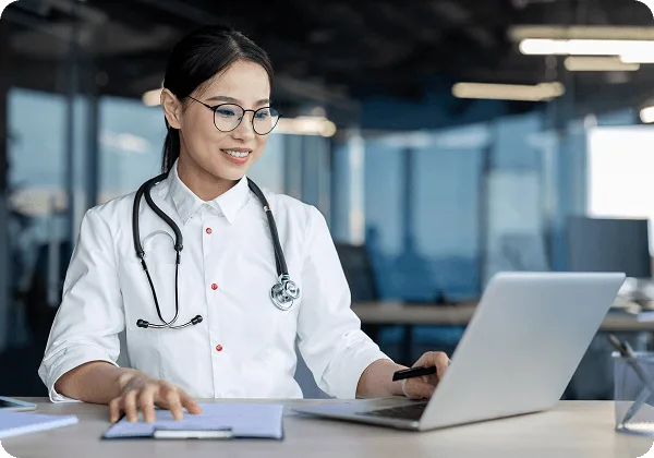patient-statements image 60610 webp Smiling woman in white coat with stethoscope and glasses working on a laptop at a desk