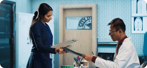 Female staff member in navy scrubs handing documents to a seated doctor in a white coat in a clinic office