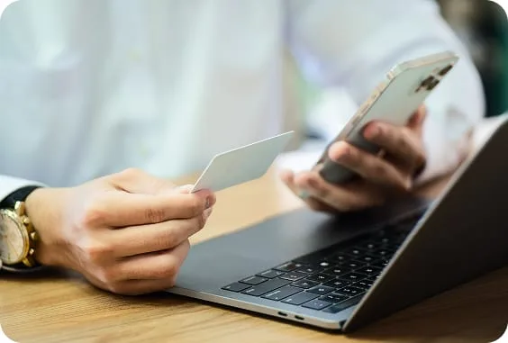 Hand holding a payment card and smartphone above an open laptop on a wooden desk