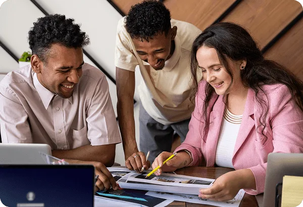 Three smiling colleagues gathered around printed documents at a table with laptops, one holding a pencil
