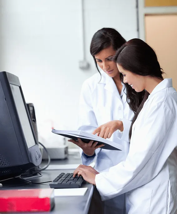 pediatric image 59492 webp Two women in white lab coats looking at an open folder beside a computer keyboard and monitor