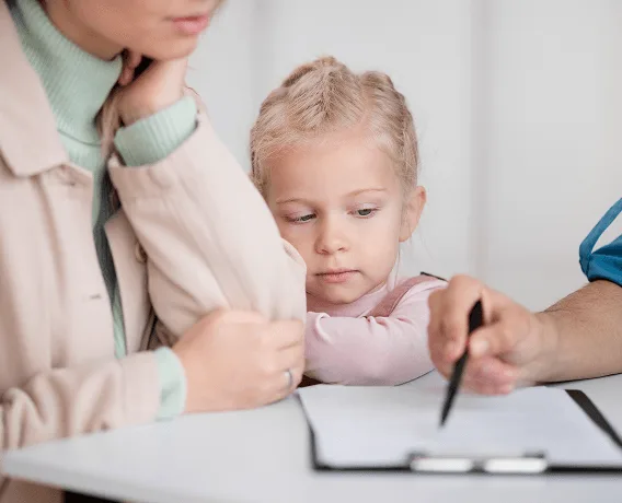 pediatric image 59494 webp Parent and young child sitting at a table while a hand points a pen at a clipboard form